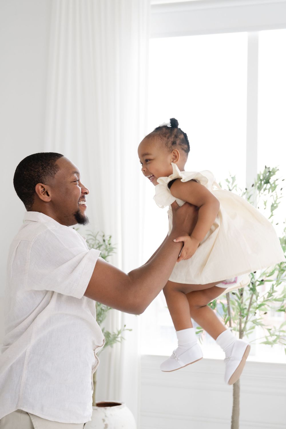 Dad with white shirt lifts a smiling toddler in a white dress. Captured by family photographer Phoebe Lee Photo from Toronto, Ontario.