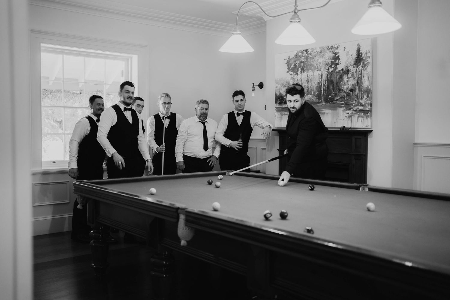 A black and white photograph shows people gathered around a pool table in a room with pendant lighting and white walls.