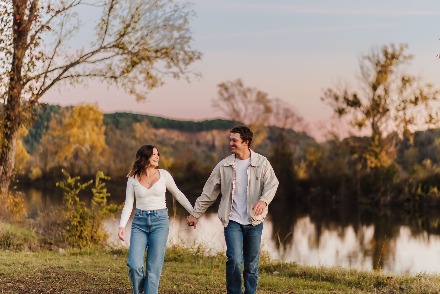 Casual couple walking hand in hand by lakeside during autumn sunset.