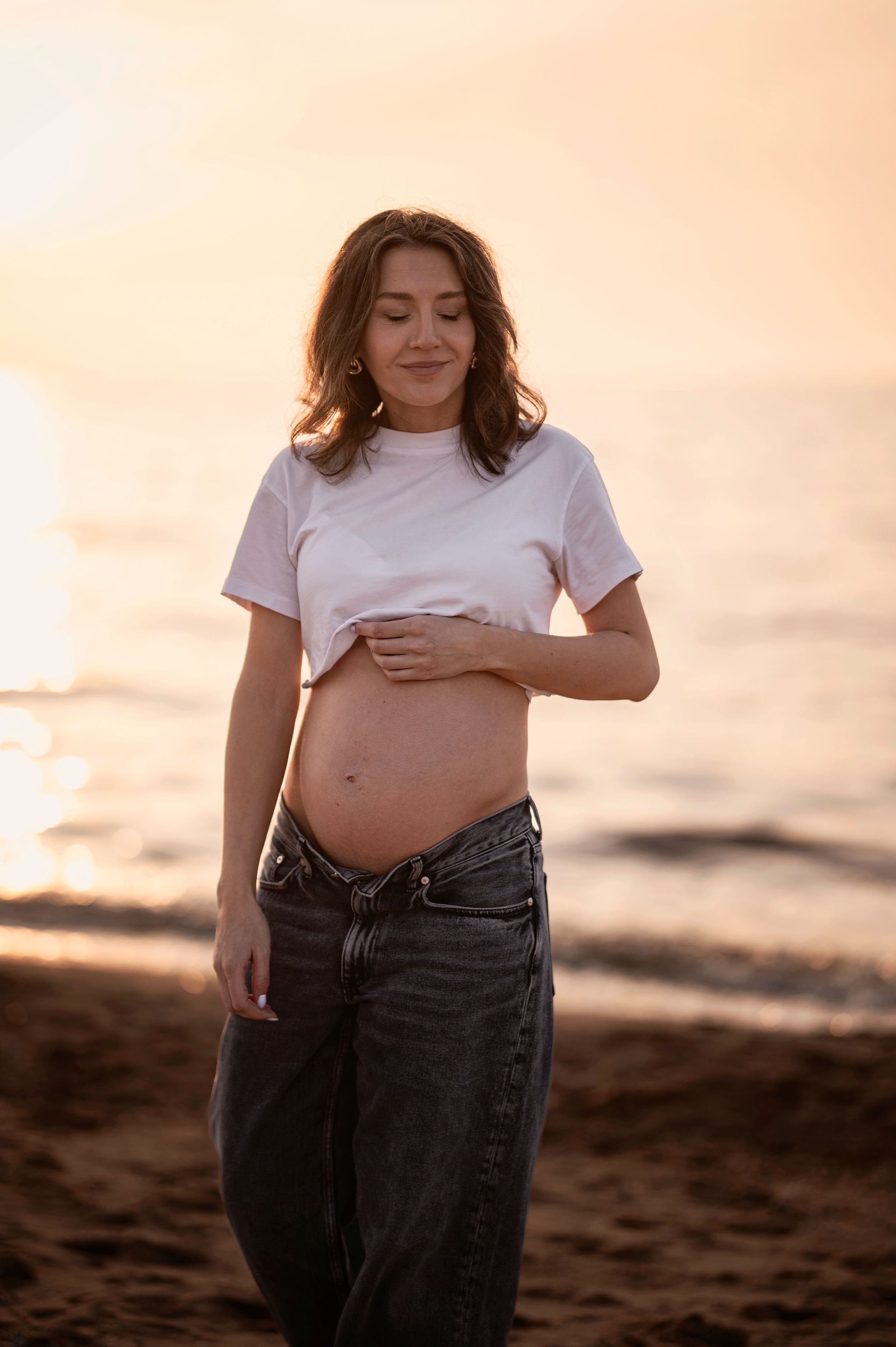 Expectant mother in white t-shirt and jeans poses on beach during golden hour sunset.