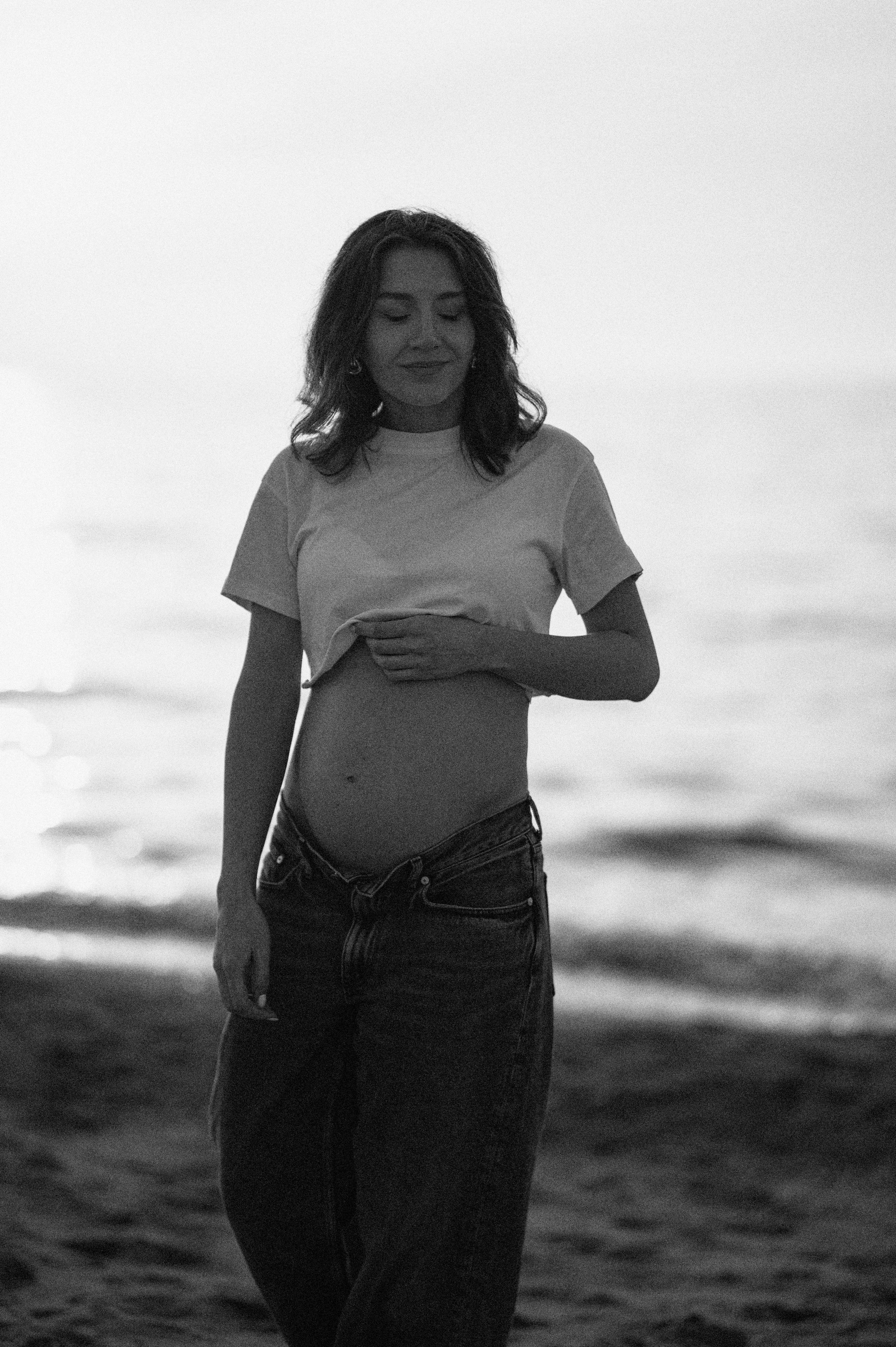 A black and white beach maternity photo showing an expectant mother holding her belly against an ocean backdrop.