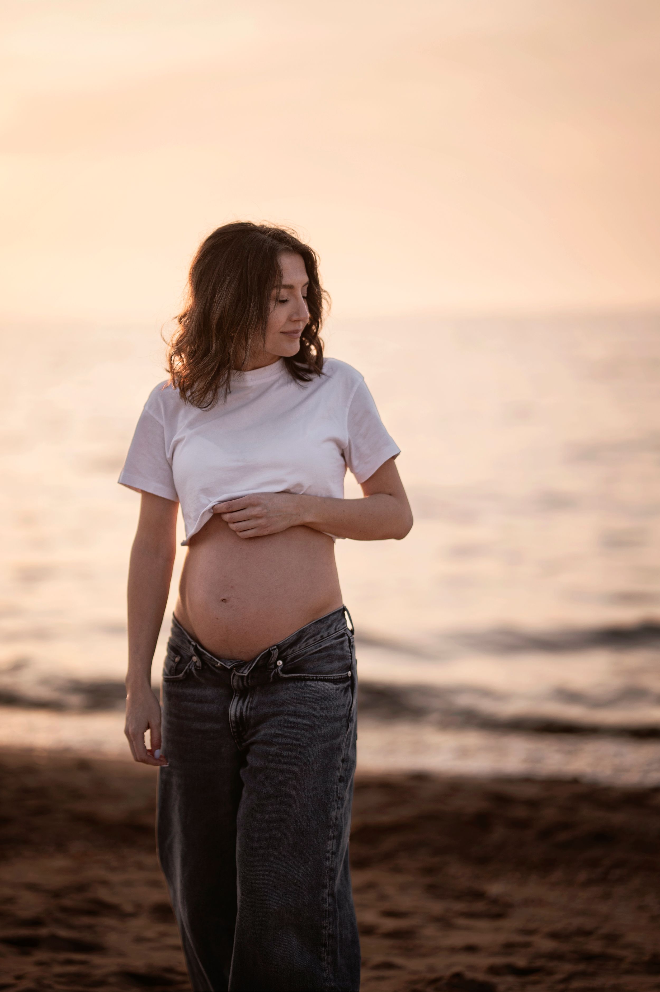 Maternity photoshoot at sunset showing pregnant woman in casual attire on beach.