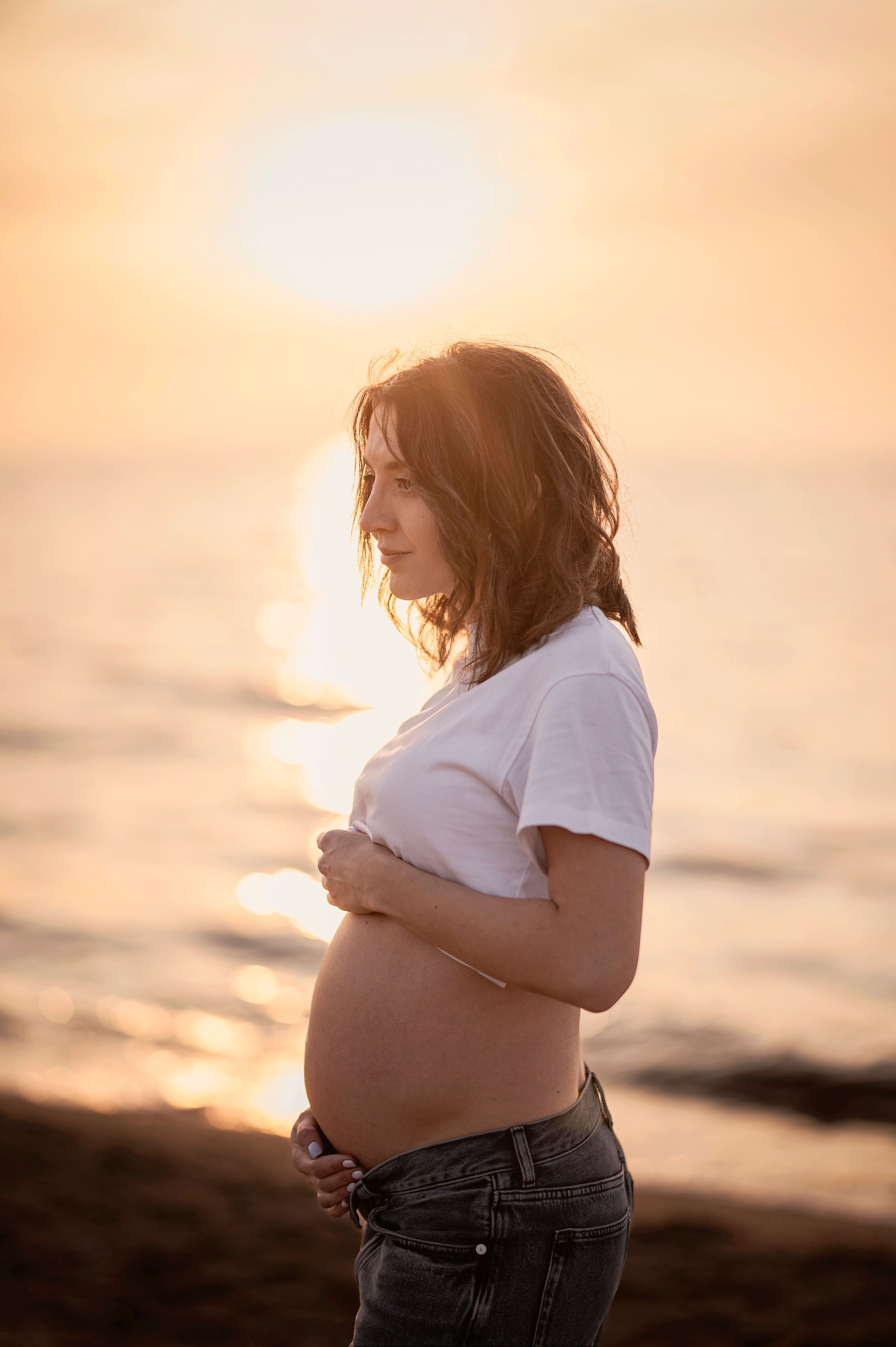 Expectant mother in white t-shirt and jeans poses at sunset on beach with gentle ocean waves in background.