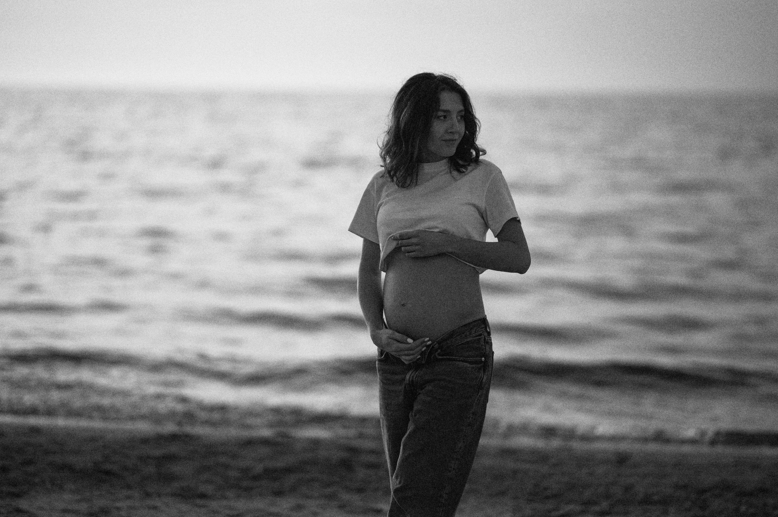 Black and white photo of a person standing on a beach at sunset with the ocean in the background.