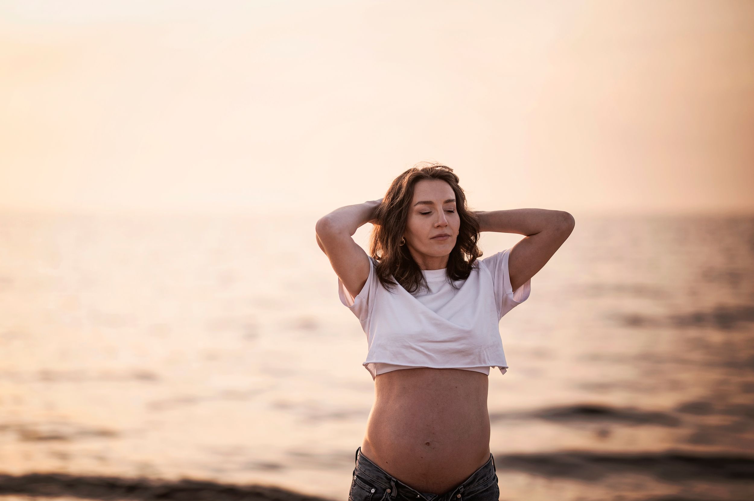 Woman in white crop top poses with hands behind head at sunset on beach with orange sky and ocean in background.