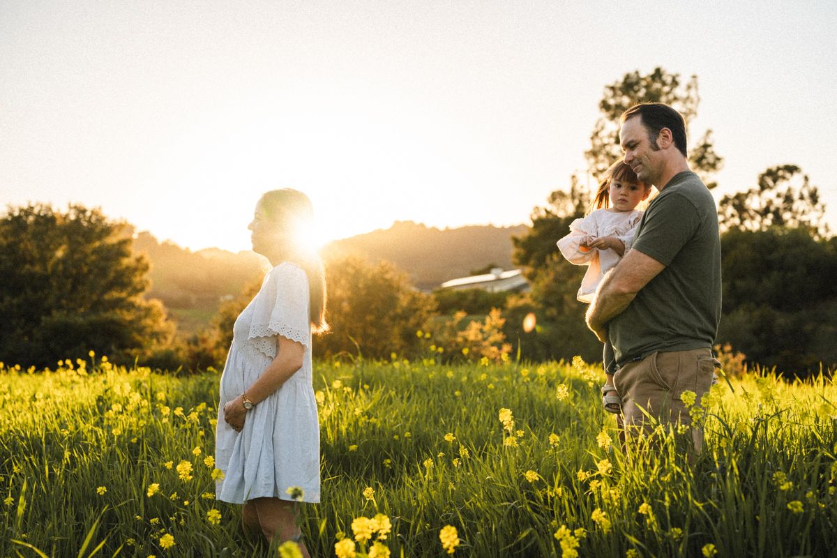 A couple stands together in a sunlit meadow filled with yellow wildflowers during golden hour.