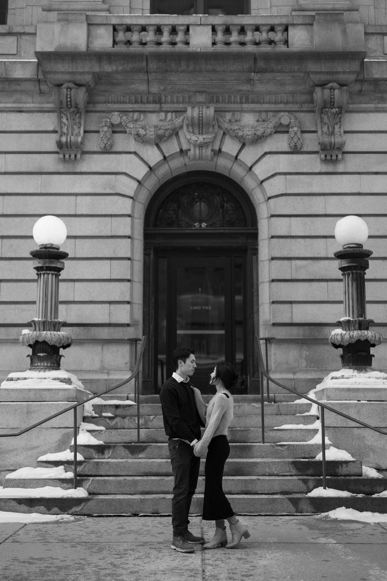A romantic black and white photo on the steps of a grand historic building with ornate lamp posts and arched entrance.
