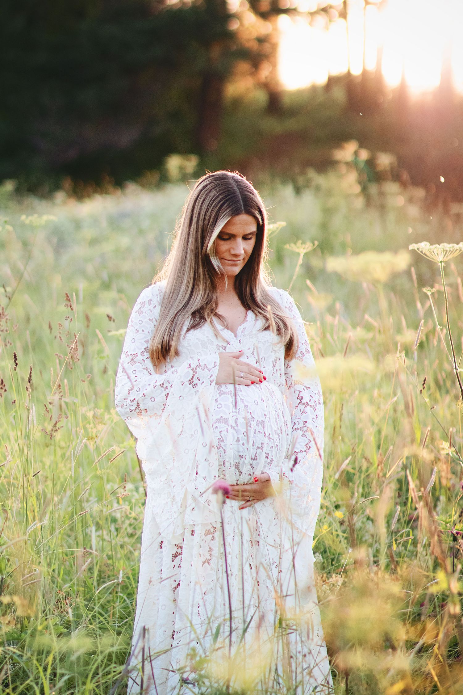 Pregnant woman in white lace gown cradles belly in sunlit wildflower meadow.