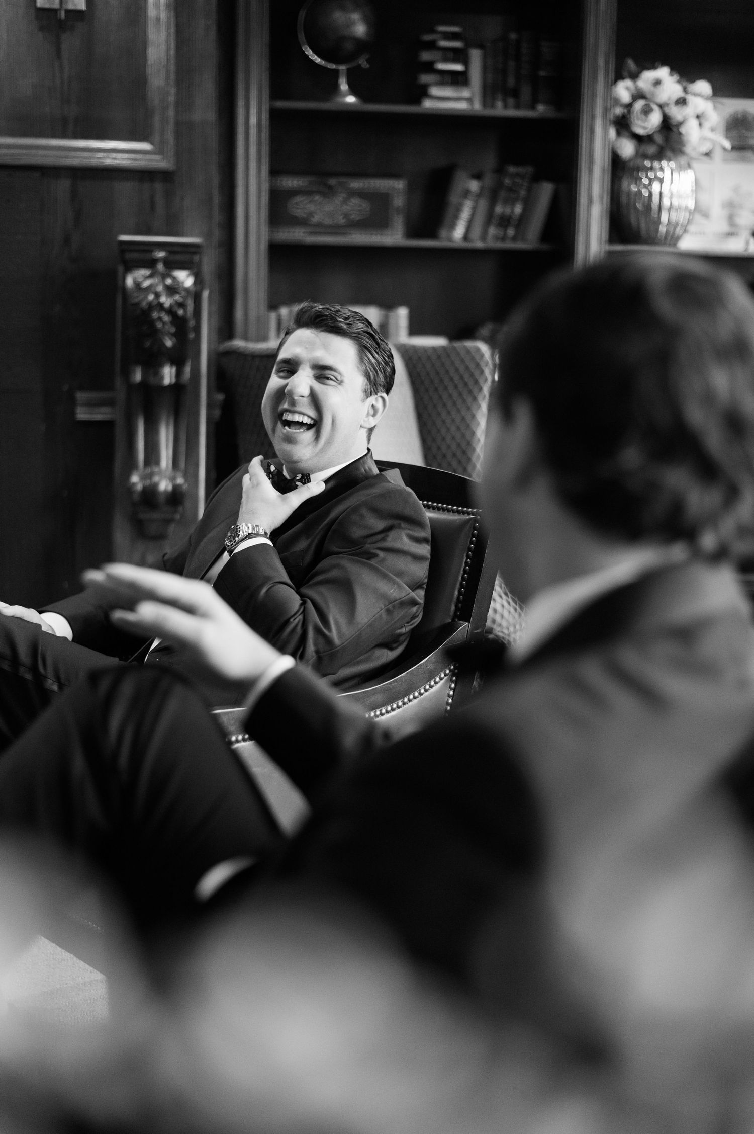 Black and white photo of someone laughing heartily while seated in an elegant armchair with bookshelves in background.