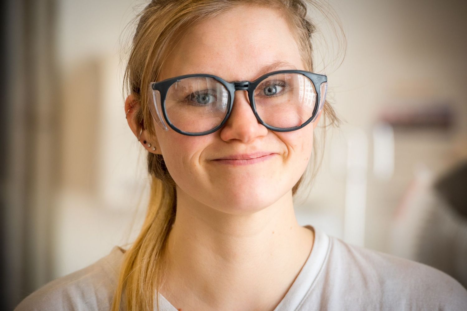 A person wearing glasses smiles warmly at the camera while wearing a white t-shirt.