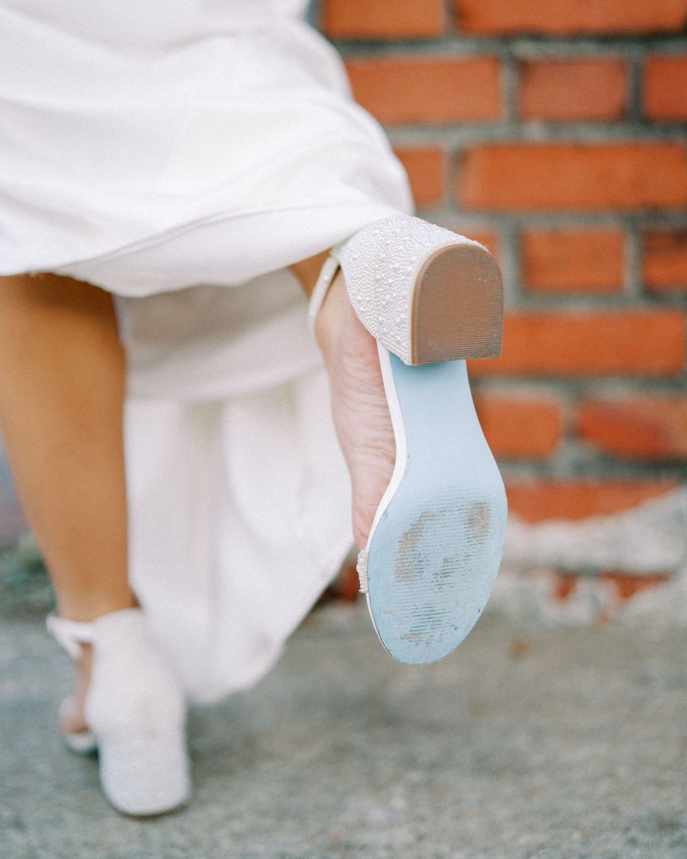 Close-up of a bride's white wedding shoe with blue sole against a brick wall background.