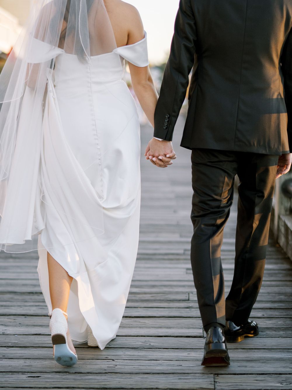 Close-up view of wedding couple walking together on wooden boardwalk showing dress and suit details.