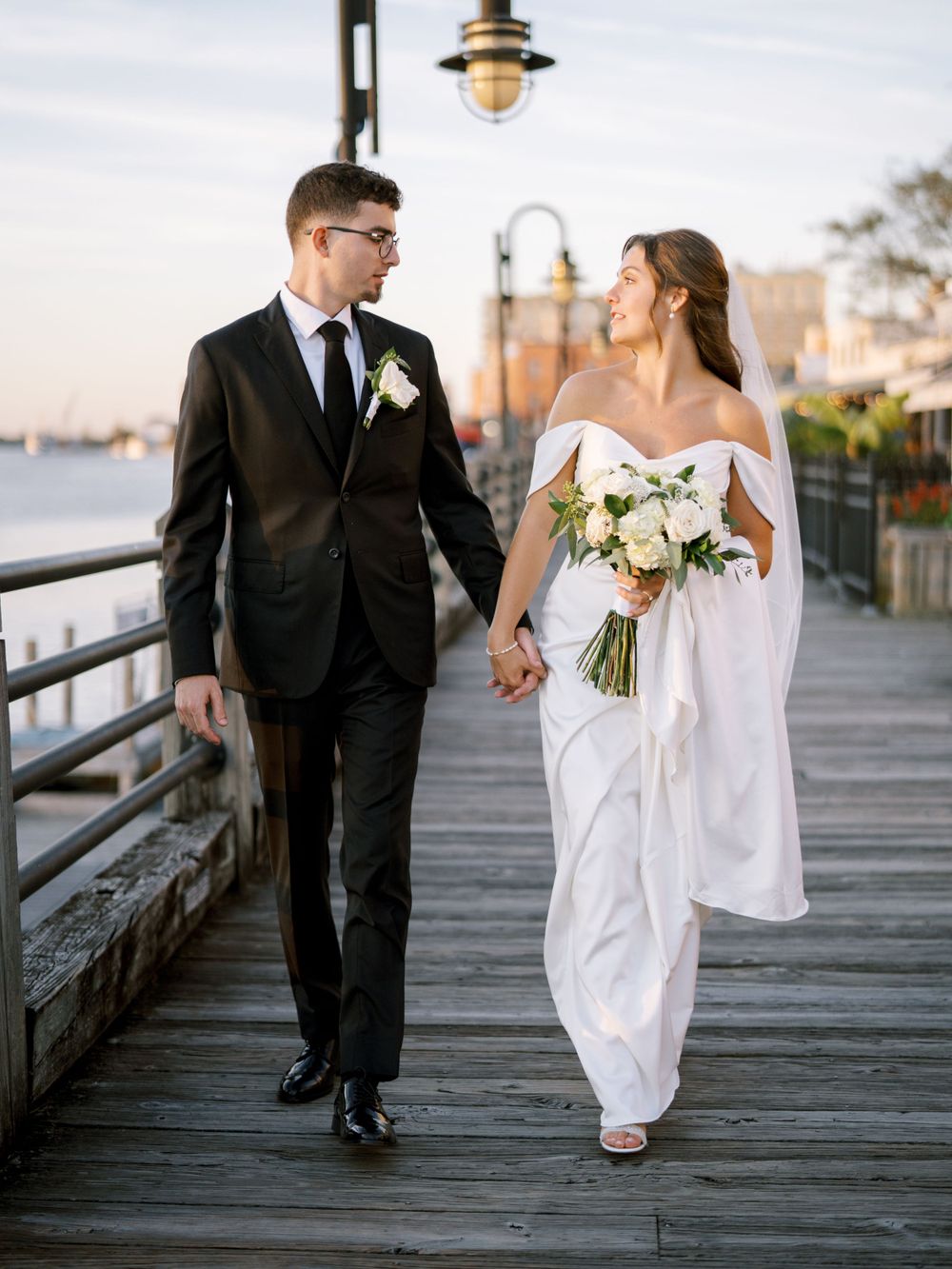 A couple in wedding attire walks on a wooden pier with vintage lampposts.