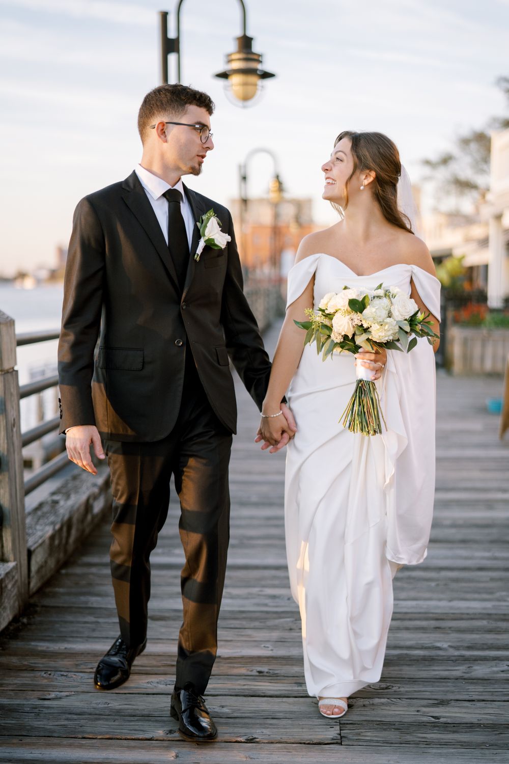 A couple walks hand in hand on a wooden pier at sunset after their wedding ceremony.