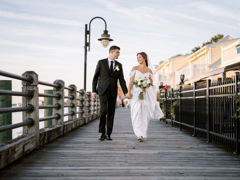 A couple strolls along a waterfront boardwalk in formal wedding attire.