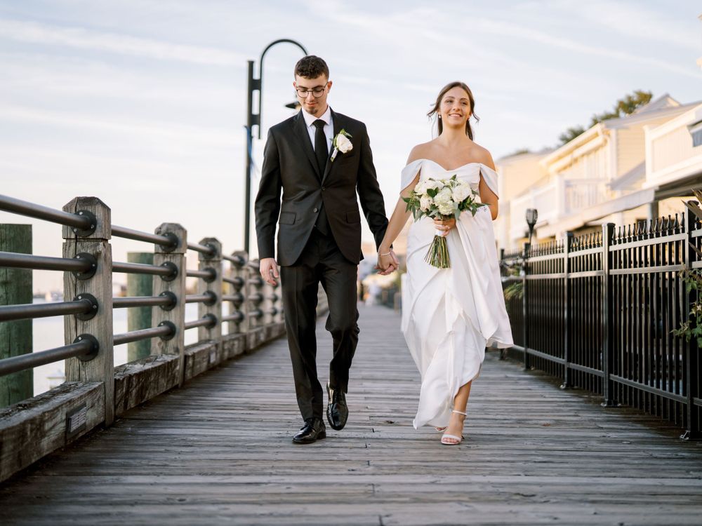 Newlyweds walk hand in hand along a wooden pier with bouquet and formal attire.