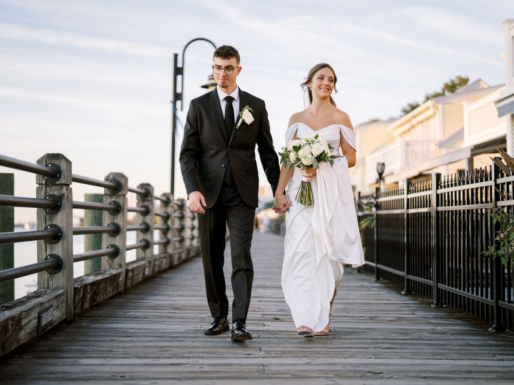 A wedding couple strolls together on a waterfront boardwalk with industrial architecture behind them.