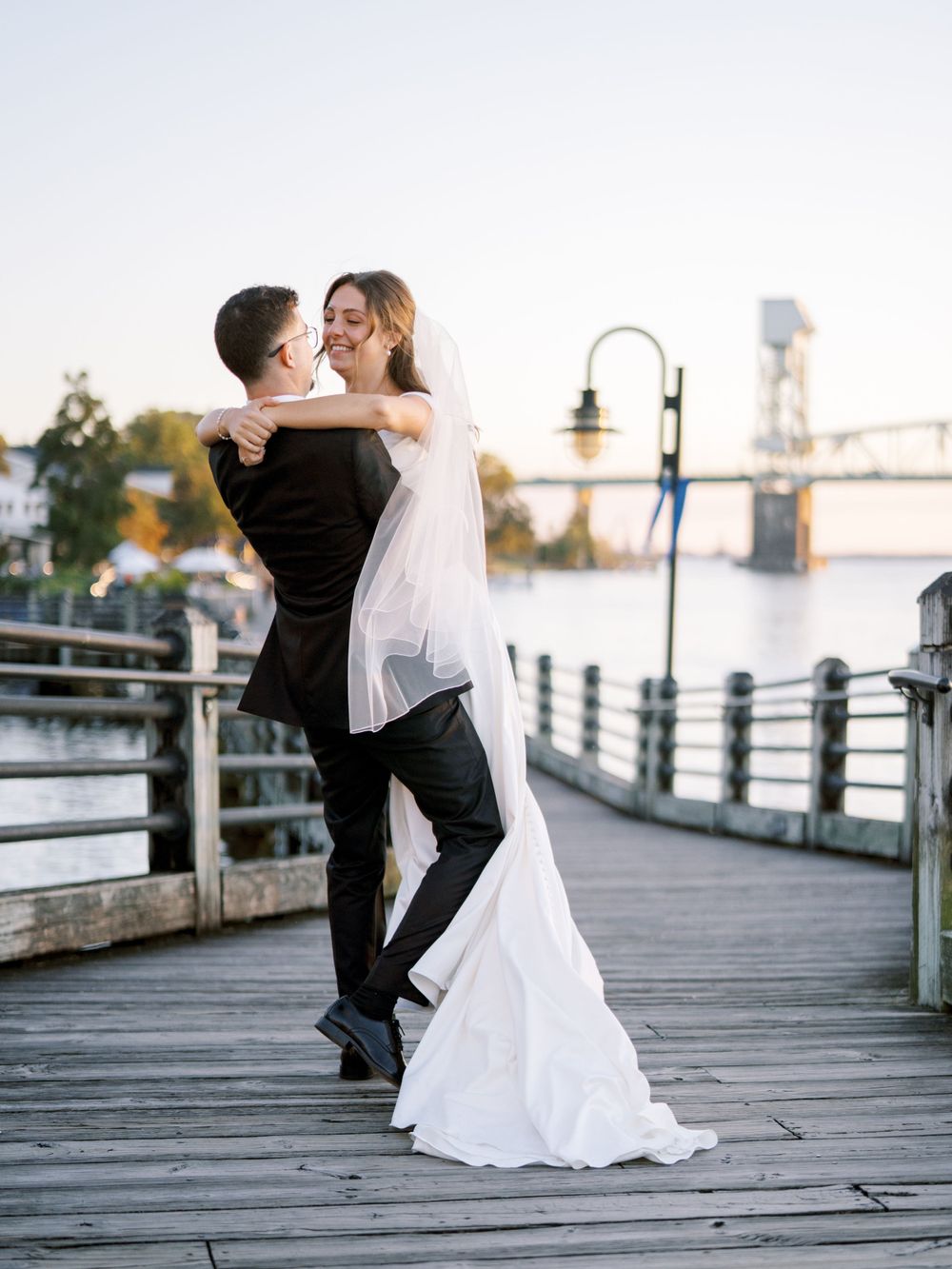 A romantic wedding couple embraces on a waterfront boardwalk at sunset with a bridge in the background.