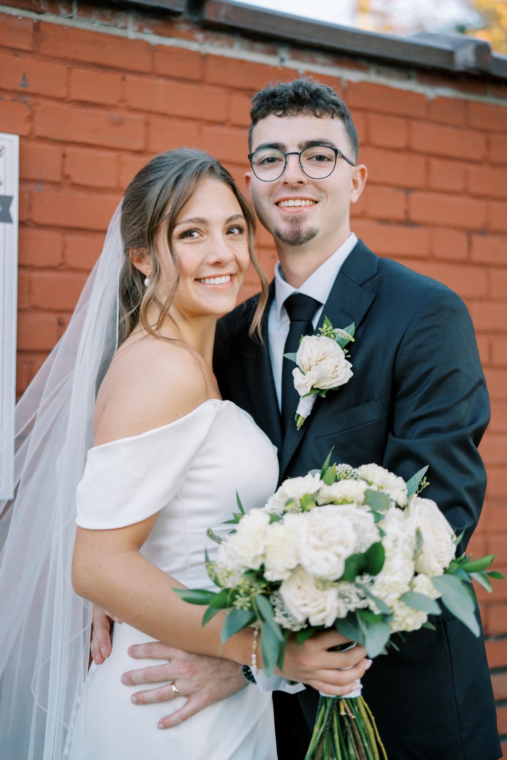 A wedding couple poses with white bouquet against a rustic brick wall background.