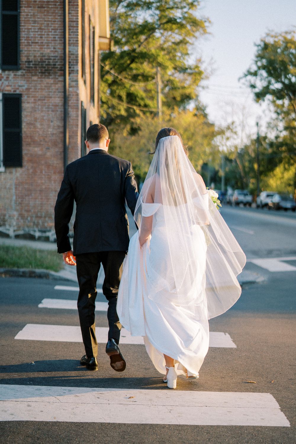 Newlyweds walking across crosswalk with bride's veil flowing in elegant black and white attire.