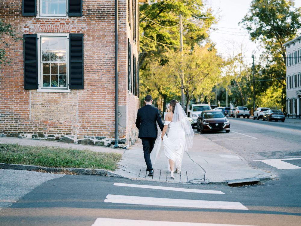 Bride and groom walking down a tree-lined street past historic brick buildings.