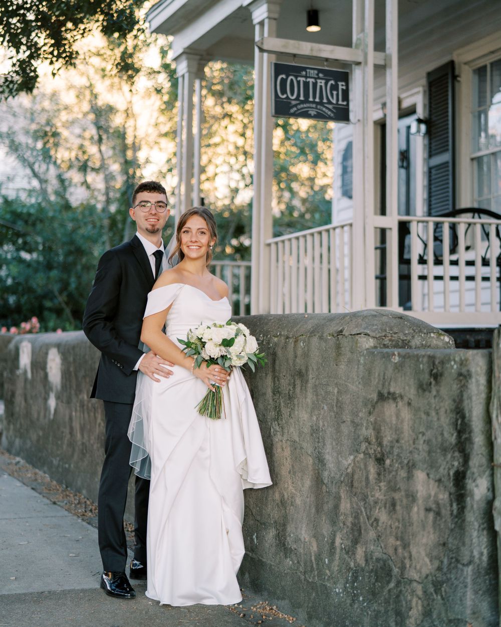 Wedding couple poses outside historic cottage building at sunset with white floral bouquet.