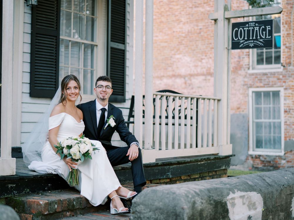 Wedding couple sitting on stone wall outside historic cottage building with white bouquet.