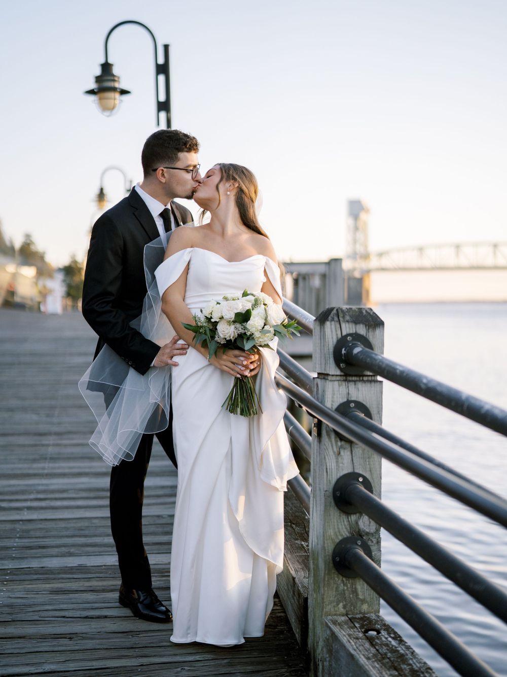 Romantic wedding portrait of couple on wooden pier with waterfront and lamp post at sunset.