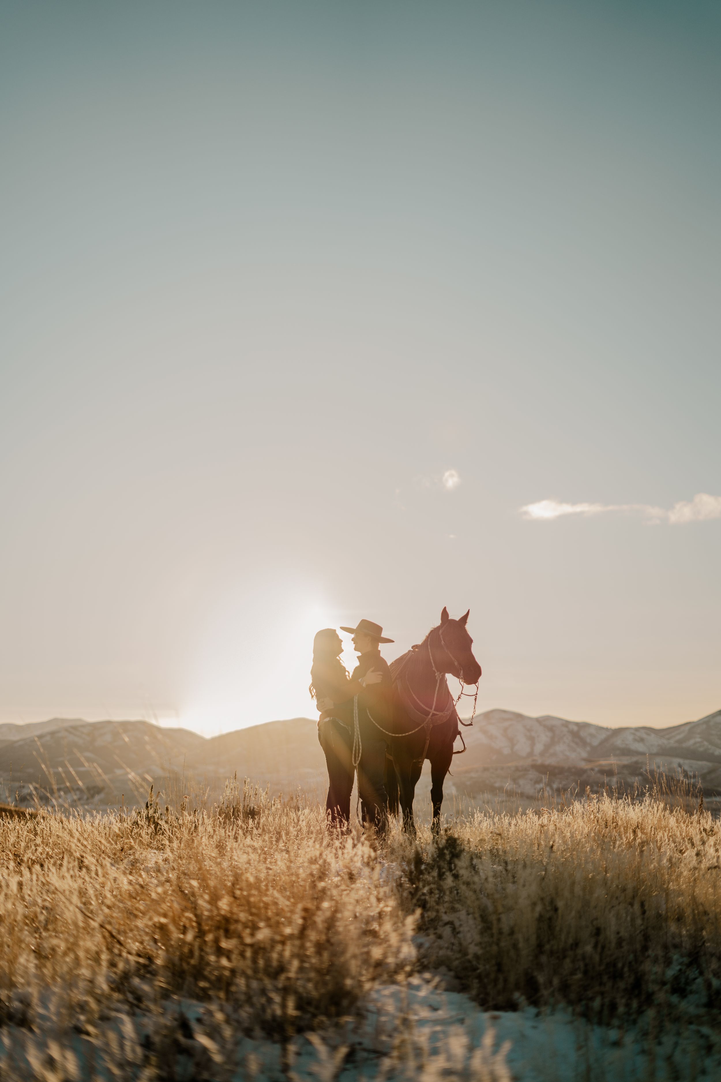 A young couple gazing into each others eyes with a horse beside them and the mountains and sunset in the background