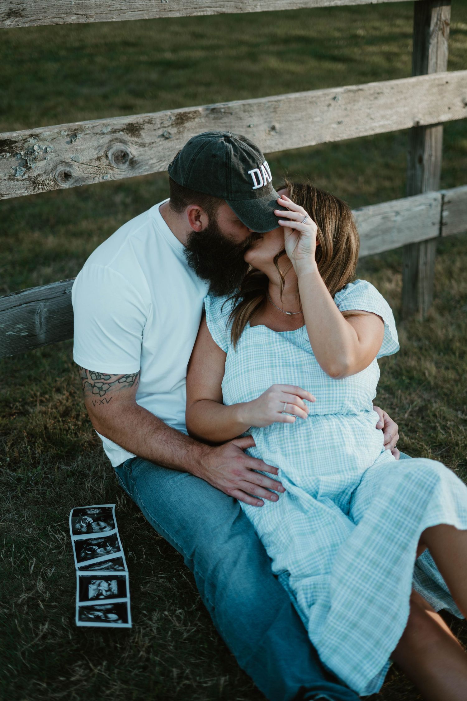 Intimate couple sitting by rustic fence with ultrasound photos laid out on ground during maternity photoshoot.