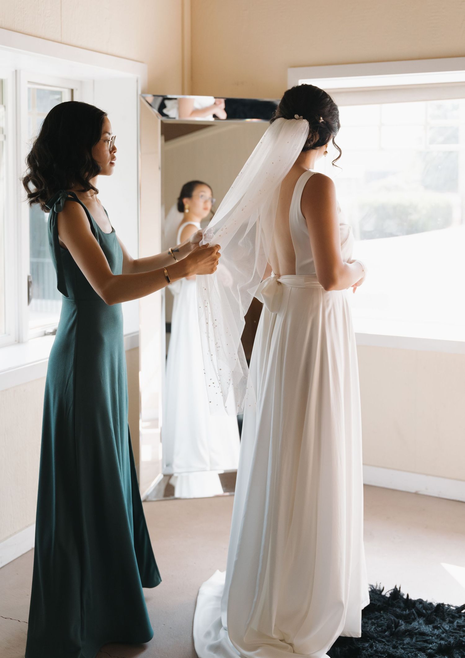 Bridesmaid in teal dress helps adjust the wedding veil in a sunlit room.
