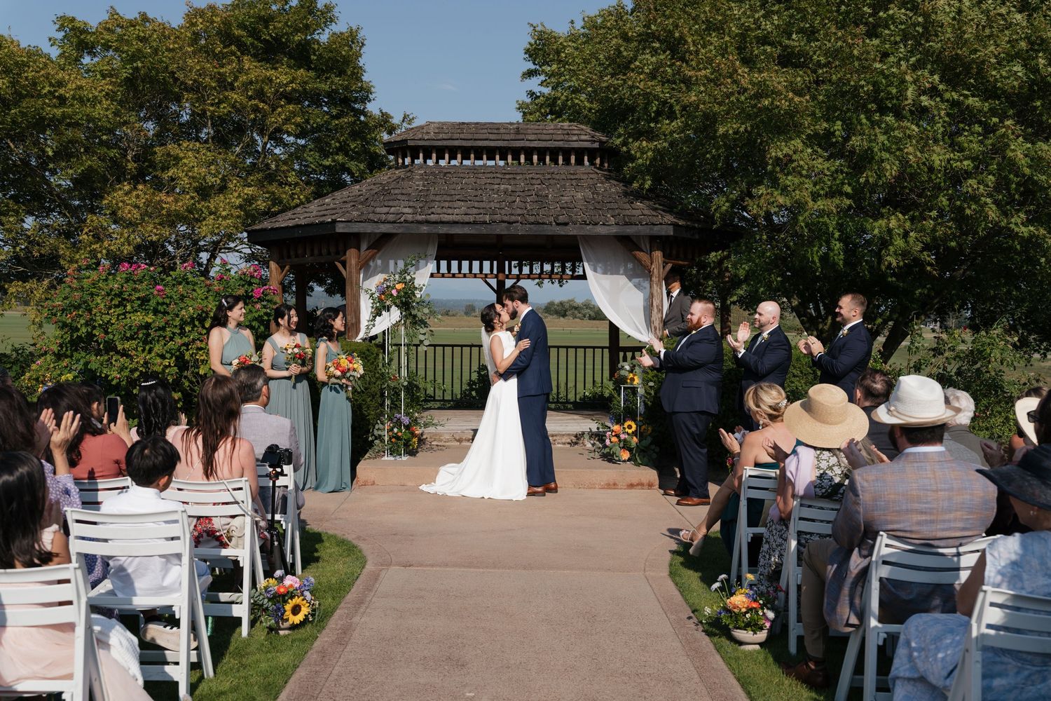 Outdoor wedding ceremony under a gazebo with guests seated on white chairs.