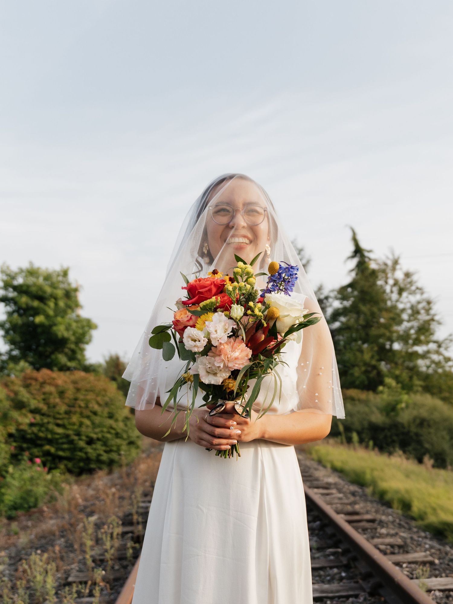 Bride holding a vibrant bouquet of mixed flowers poses on train tracks.