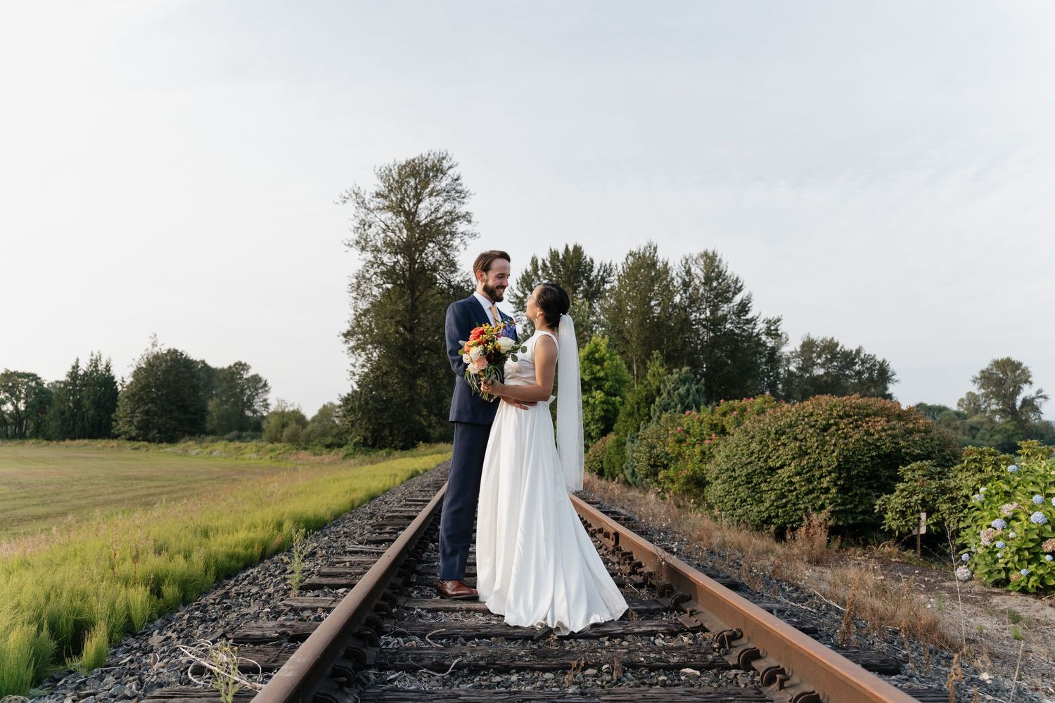 Newlyweds embrace while standing on railroad tracks surrounded by greenery.