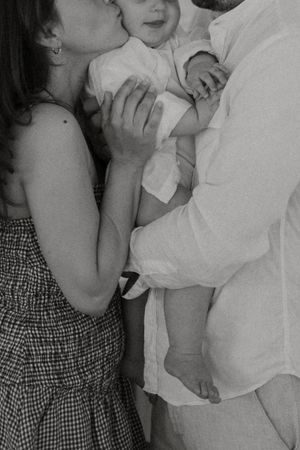 Black and white intimate family moment showing a baby being held by parents wearing white and patterned clothing.
