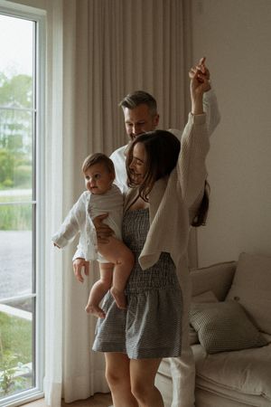 A joyful family dances together in a sunlit living room beside large windows and neutral colored curtains.
