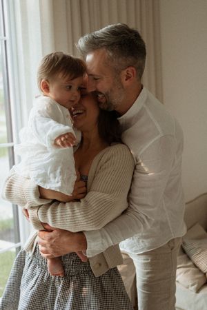 A tender family moment captured indoors as they share an intimate embrace in soft natural light by the window.