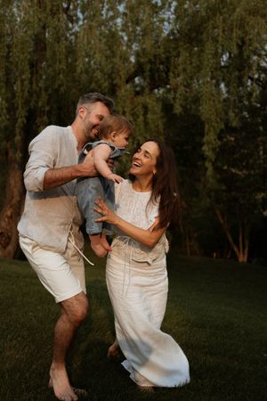 Family of three sharing joyful moment outdoors in white attire against wooded background at golden hour.