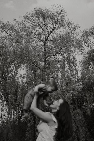 Black and white portrait of a mother lifting her child playfully against a backdrop of tall trees.