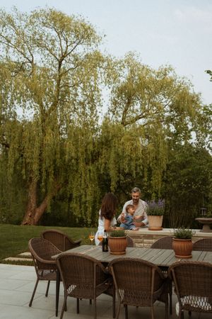 People enjoying an outdoor patio setting with wicker furniture and potted plants under a weeping willow tree backdrop.
