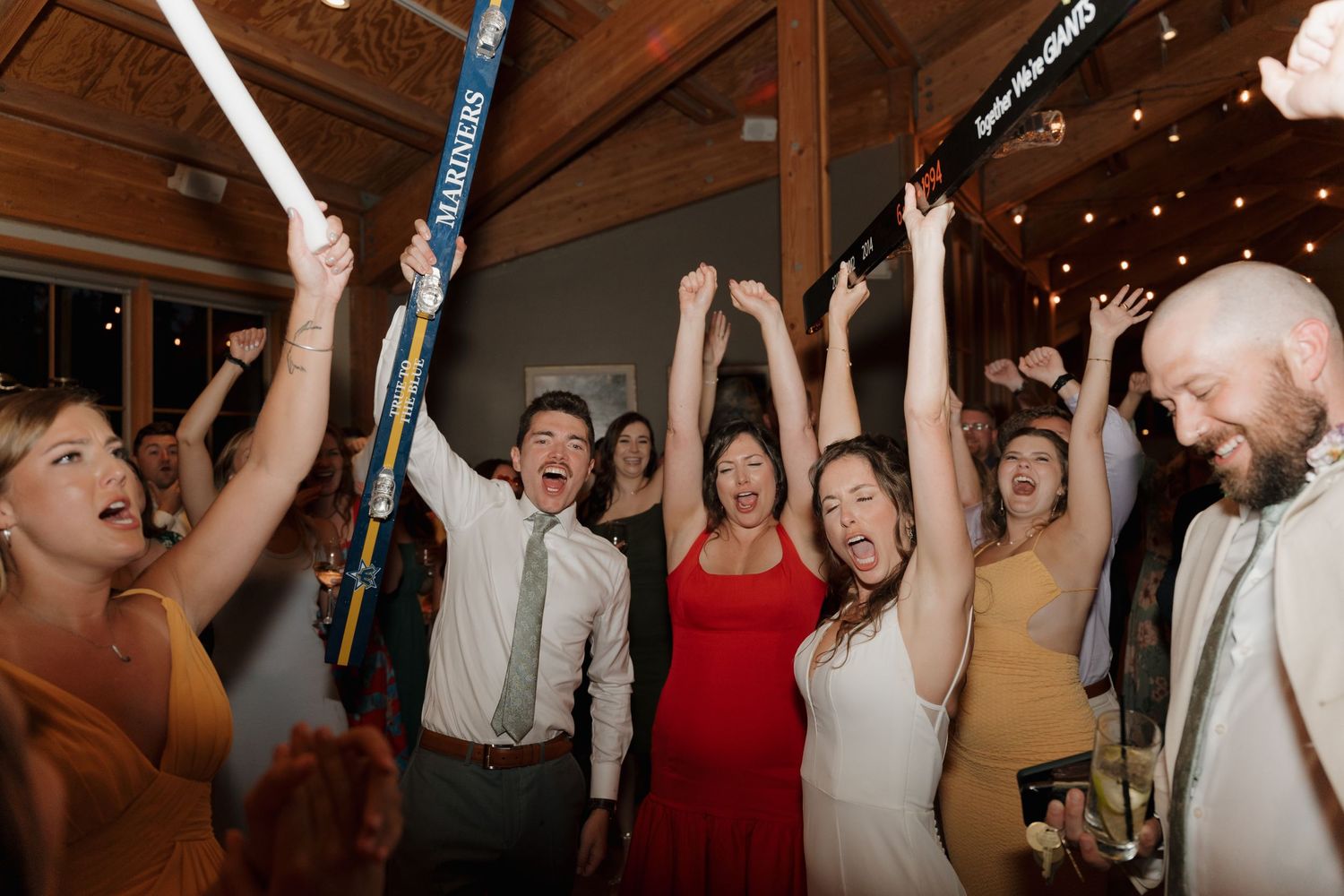 Wedding guests celebrate joyfully with raised arms at a lively indoor reception.