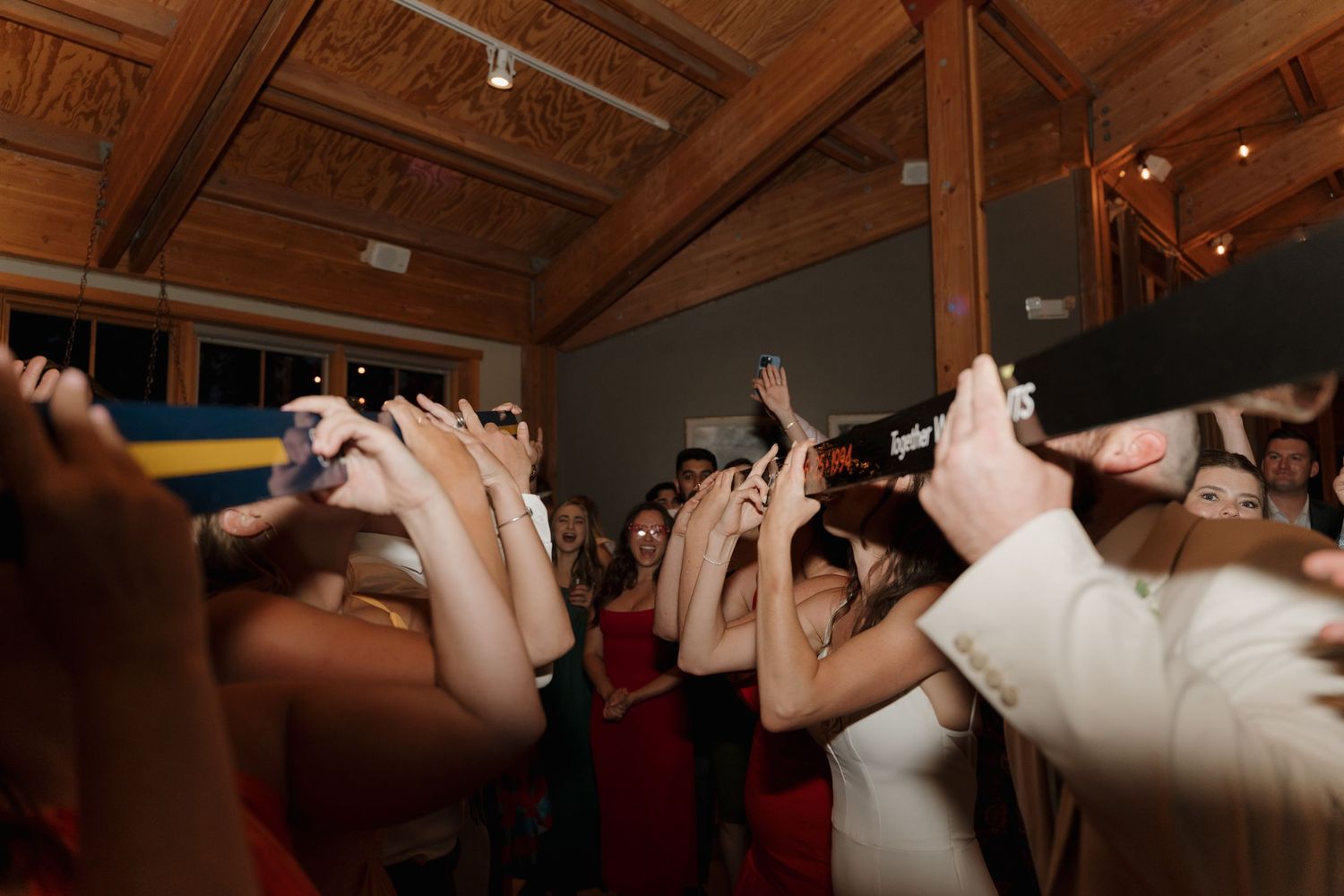 Energetic wedding reception crowd cheers with hands in the air under wooden beams.
