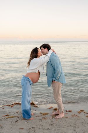 A couple embraces on a beach at sunset with gentle waves and sand in the background.