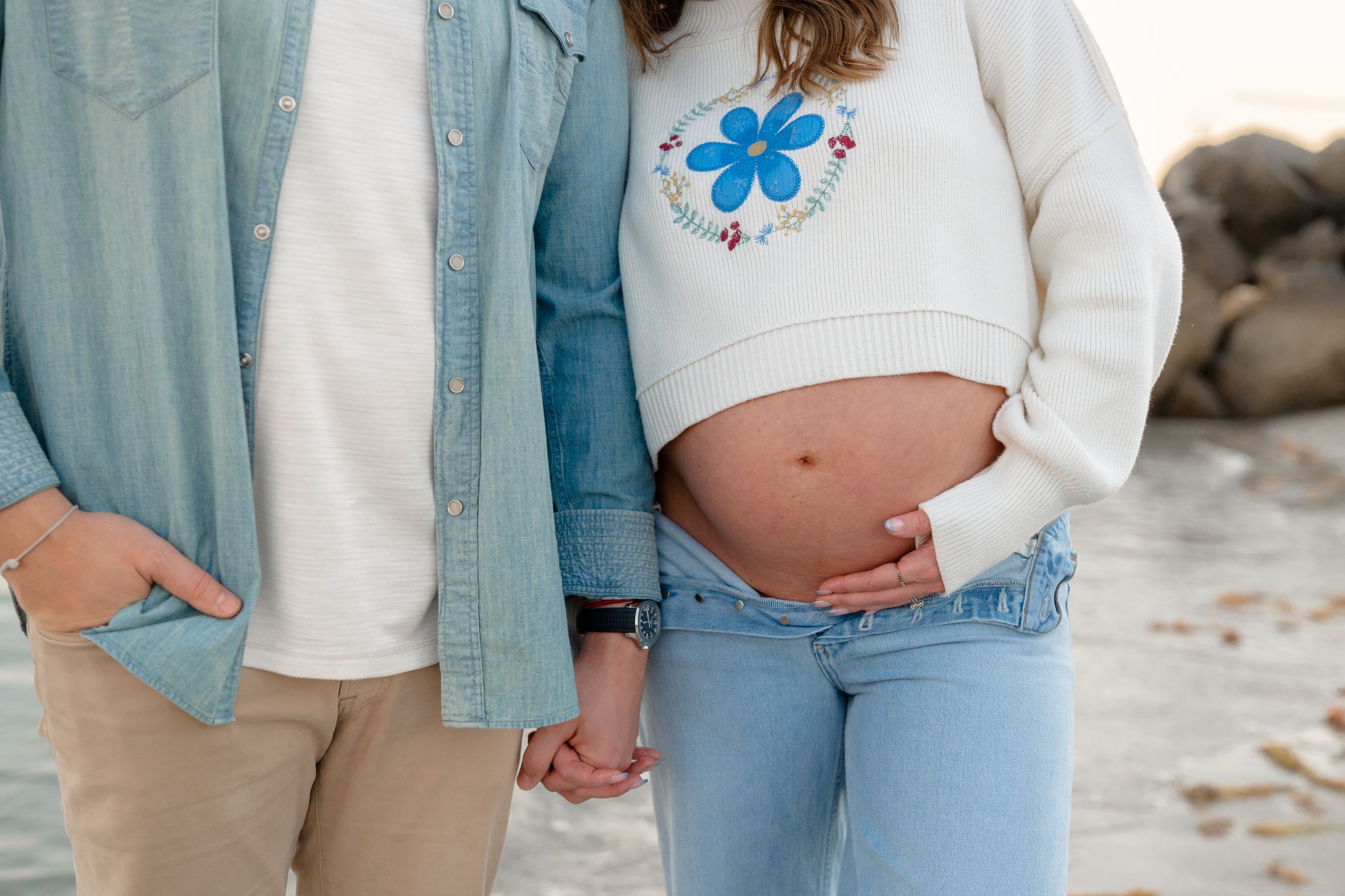 A couple in matching denim and white outfits holds hands while walking on a beach during a maternity photoshoot.