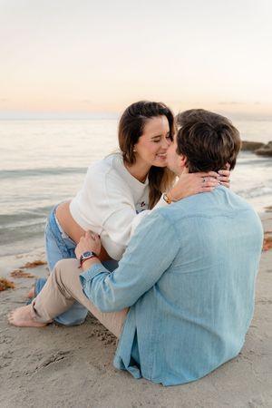 A couple in casual attire shares an intimate moment while sitting together on a sandy beach at sunset.