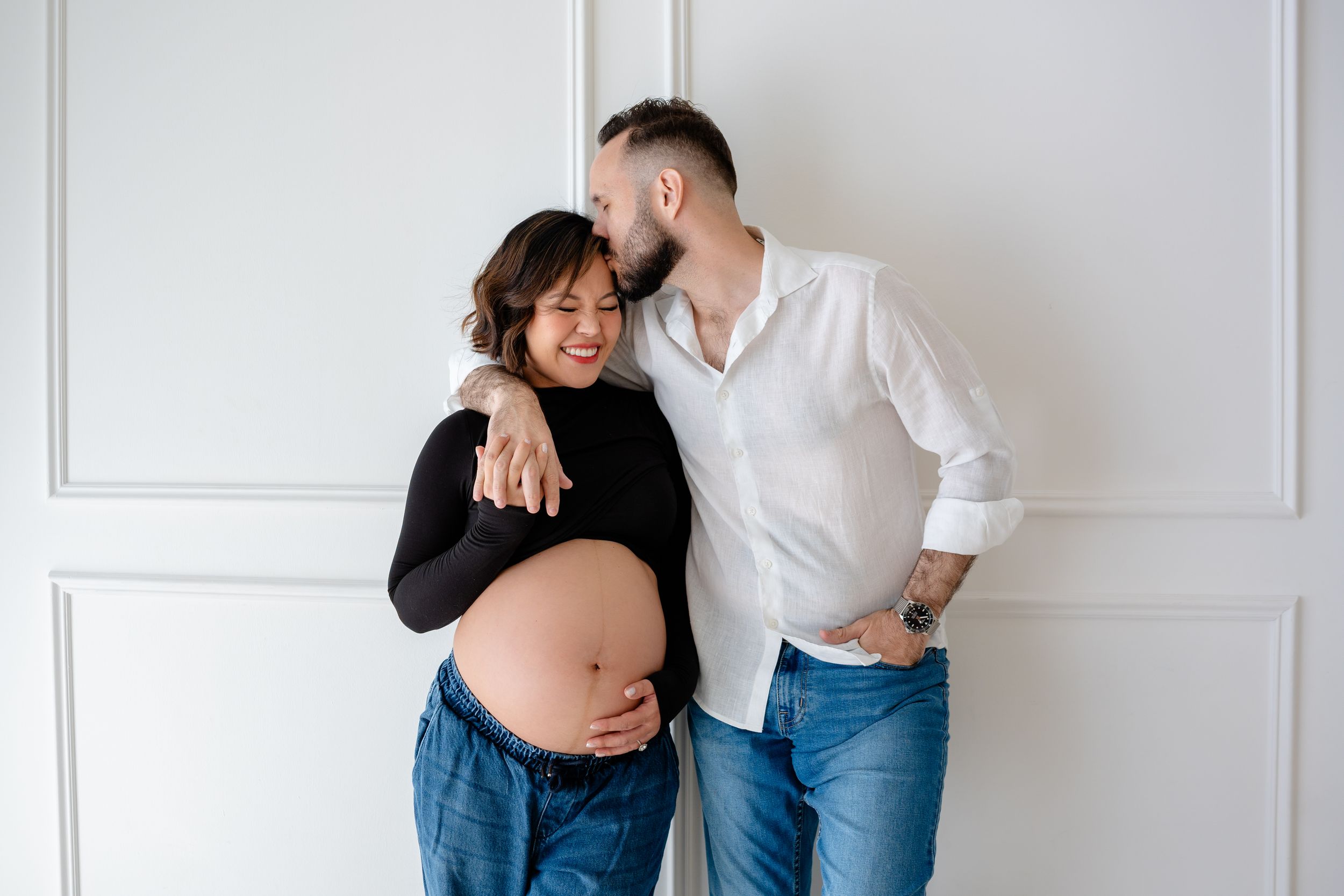 A couple in casual attire shares intimate moments against a white wall in a romantic photoshoot.