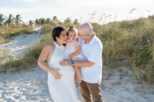 Family wearing white and tan outfits embraces while standing on sandy beach path with palm trees in background.