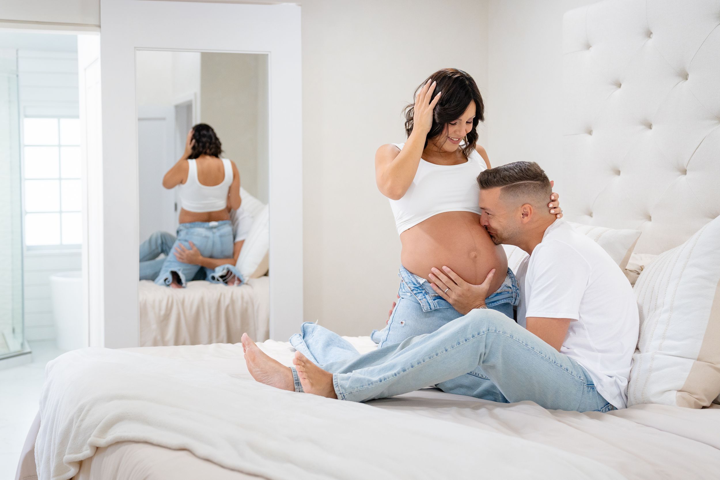 Intimate pregnancy photography session on a white bed with mirror reflection in a bright bedroom setting.