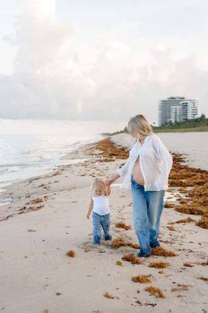 Parent and child walk together on a beach picking up seaweed during a cloudy afternoon.