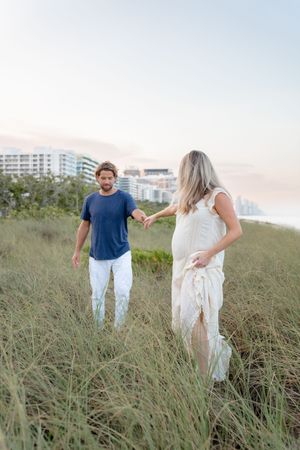Couple walking and playing together through tall grass on a beach dune with coastal buildings in the background.